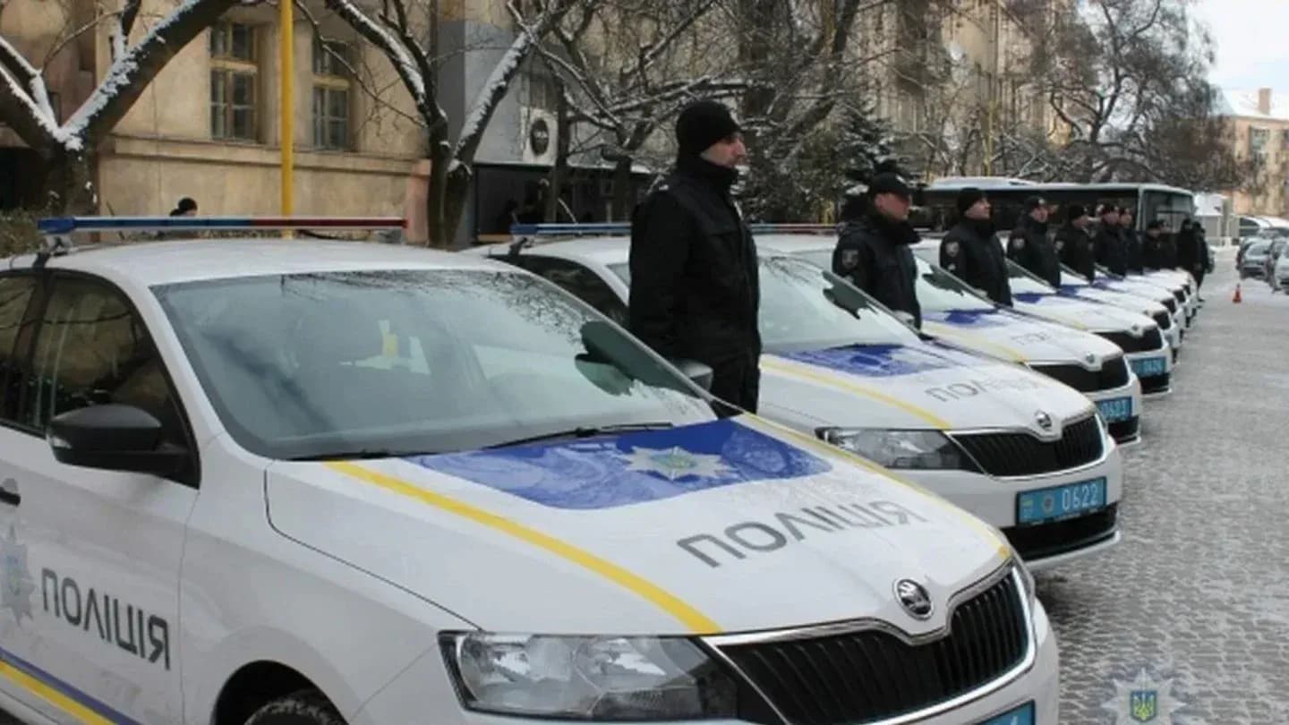 Ряд белых автомобилей Škoda с сине-жёлтой окраской и кириллической надписью «ПОЛІЦІЯ» припаркован вдоль заснеженной брусчатки, рядом с машинами стоят люди в тёмной зимней форме и фуражках, возле голых деревьев и зданий.
